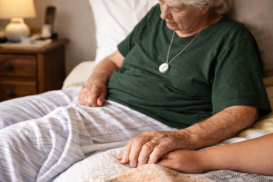 Close-up of an older woman in a dark green shirt sitting on a bed, holding another person's hand